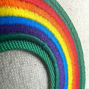 May include: Embroidered rainbow patch with a green border. The rainbow features vibrant colors: red, orange, yellow, green, blue, and purple. The patch is likely for clothing or accessories.
