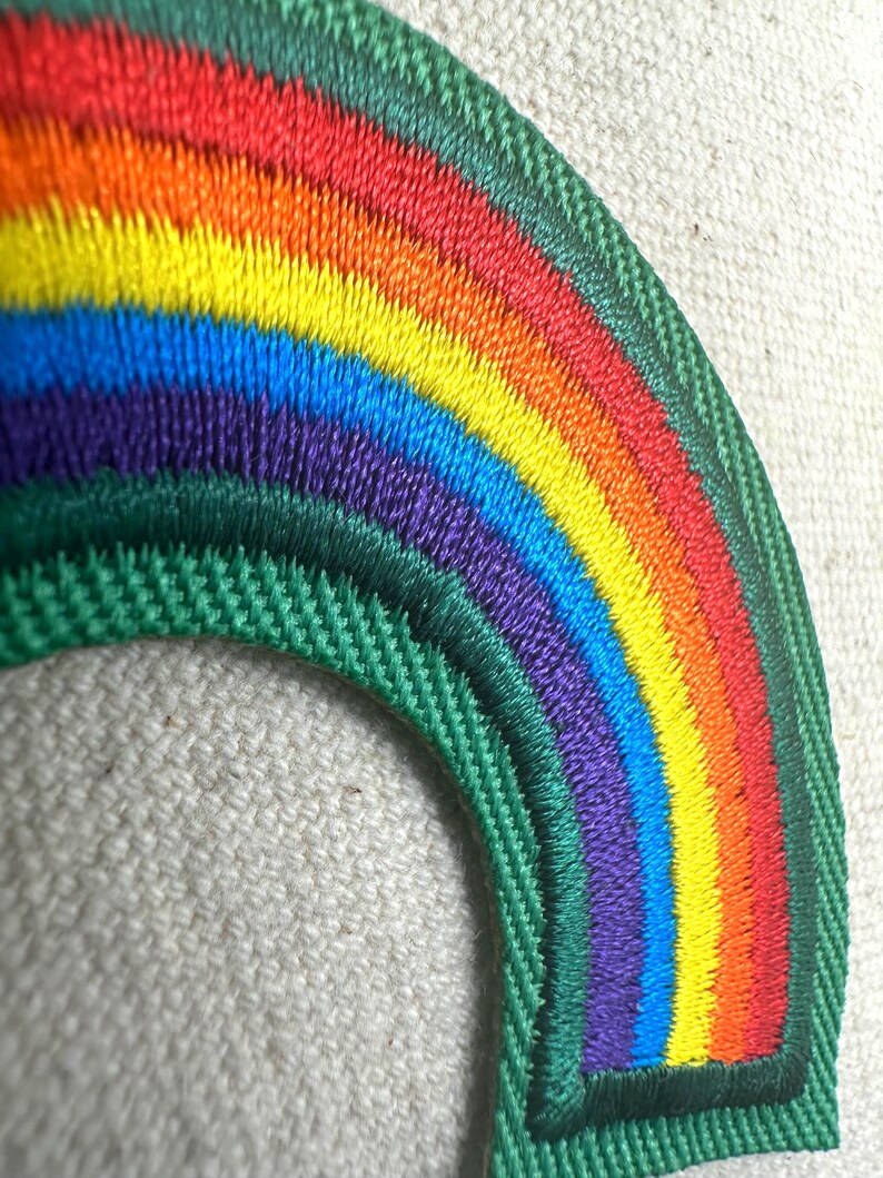 May include: Embroidered rainbow patch with a green border. The rainbow features vibrant colors: red, orange, yellow, green, blue, and purple. The patch is likely for clothing or accessories.