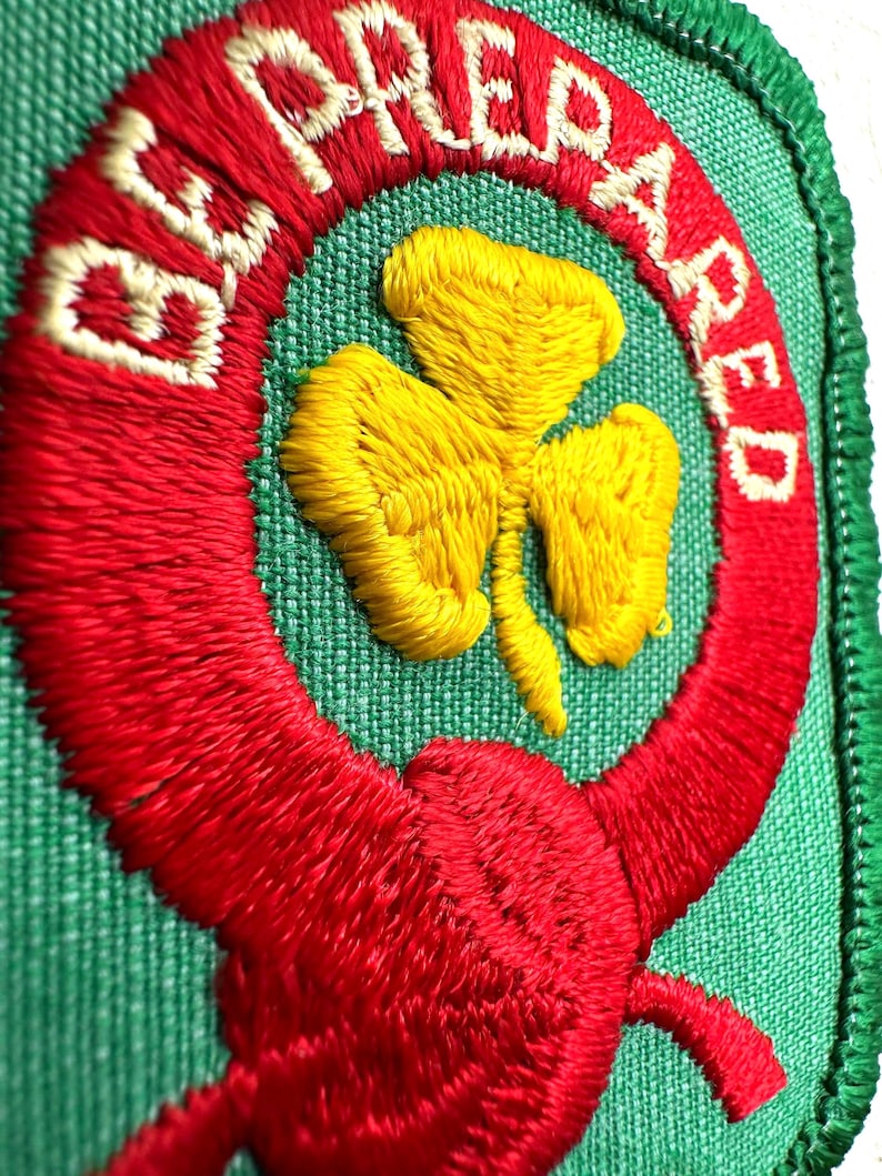 May include: Embroidered patch featuring the text "BE PREPARED" in white on a red ring, encircling a yellow shamrock on a green backdrop. The patch, with a green border, is likely for a scouting uniform or similar organisation.