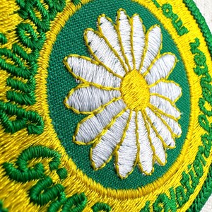 Puede incluir: Parche bordado con una margarita blanca y centro amarillo sobre fondo verde. El parche est&aacute; bordeado por un anillo amarillo y un anillo exterior verde con las palabras "Scout Squad" en cursiva. El parche es redondo.