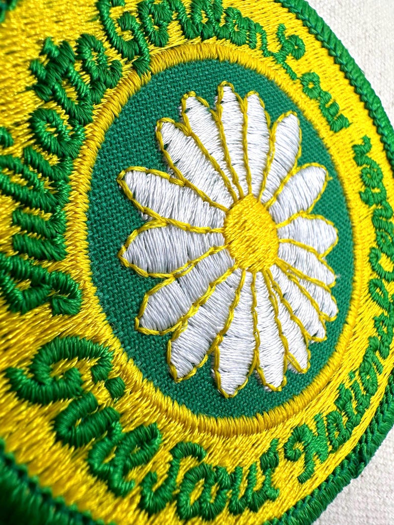 Puede incluir: Parche bordado con una margarita blanca y centro amarillo sobre fondo verde. El parche est&aacute; bordeado por un anillo amarillo y un anillo exterior verde con las palabras "Scout Squad" en cursiva. El parche es redondo.