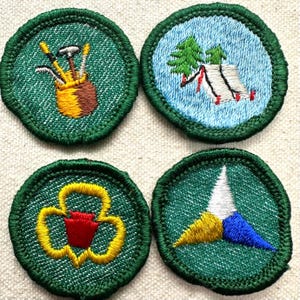 May include: Four embroidered patches with green borders. The patches feature a golf bag with clubs, a tent and trees, a trefoil, and a compass design. The patches are on a textured, off-white surface.