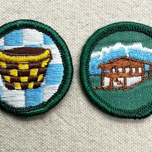 May include: Two embroidered patches with green borders. One features a brown and yellow checkered basket on a blue and white checkered background. The other depicts a brown cabin with white accents against a backdrop of blue mountains.