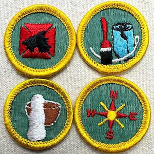 May include: Four circular patches with green centers and yellow borders. The patches feature various embroidered designs: a red and black emblem, a red brush and blue object, a white and brown object, and a compass rose. These patches are likely for a youth organization.