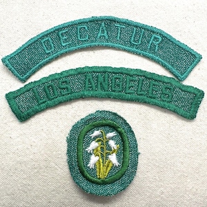 May include: Three green embroidered patches. Two are arched, with the text "DECATUR" and "LOS ANGELES". The third is a circular patch with a green border, featuring white flowers with yellow centers.