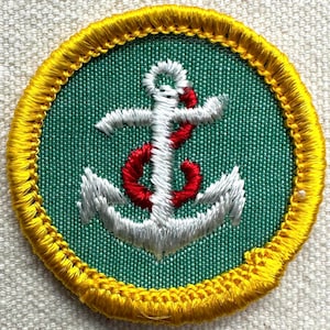 May include: Circular embroidered patch with a white and red anchor design on a green background. The patch is edged with yellow stitching. Suitable for sewing onto garments or bags.
