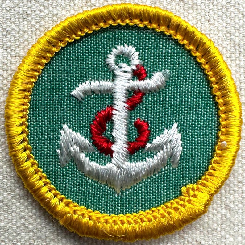 May include: Circular embroidered patch with a white and red anchor design on a green background. The patch is edged with yellow stitching. Suitable for sewing onto garments or bags.