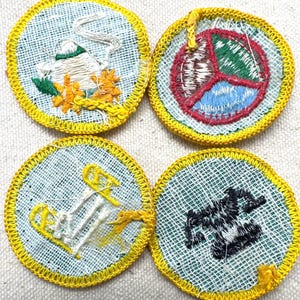 May include: Four round embroidered patches with yellow borders. The patches feature various designs: a bird, a peace symbol, an anchor, and a stylized object. The patches are on a light blue background.