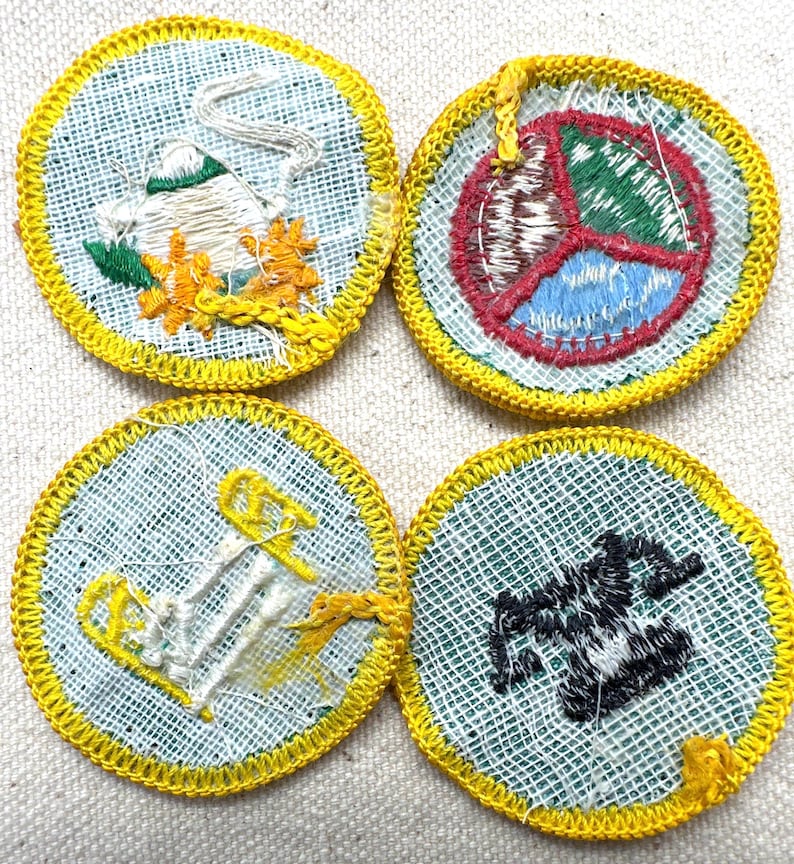 May include: Four round embroidered patches with yellow borders. The patches feature various designs: a bird, a peace symbol, an anchor, and a stylized object. The patches are on a light blue background.