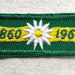 May include: Embroidered patch with a green background and a yellow banner displaying the numbers "1860" and "1960". A white and yellow flower is centered on the patch. The patch has a green border.