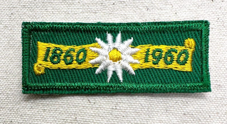 May include: Embroidered patch with a green background and a yellow banner displaying the numbers "1860" and "1960". A white and yellow flower is centered on the patch. The patch has a green border.