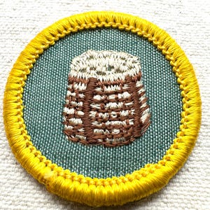 May include: Embroidered patch featuring a brown and white basket design on a teal background, encircled by a yellow border. The patch is round and appears to be made of fabric, with detailed stitching.