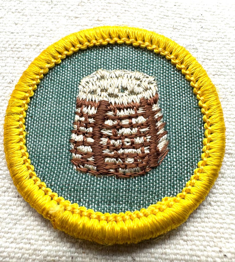 May include: Embroidered patch featuring a brown and white basket design on a teal background, encircled by a yellow border. The patch is round and appears to be made of fabric, with detailed stitching.