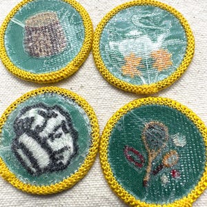 May include: Four round patches with green centers and yellow embroidered borders. The patches feature various designs, including a basket, flowers, a baseball, and a spoon with flowers. The patches are likely for a youth organization.