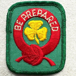 May include: A green embroidered patch featuring a red ring with the words "BE PREPARED" in white. A yellow trefoil is inside the ring. The patch has a green border.
