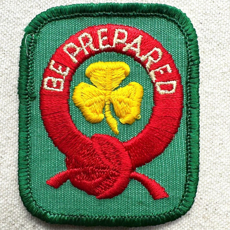 May include: A green embroidered patch featuring a red ring with the words "BE PREPARED" in white. A yellow trefoil is inside the ring. The patch has a green border.