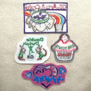 Puede incluir: Cuatro parches bordados sobre un fondo beige. Los parches incluyen un arco&iacute;ris, una magdalena, un coraz&oacute;n y dos personajes de dibujos animados. Los parches tienen varios colores, incluyendo rosa, azul, verde y blanco. Los parches tienen texto.