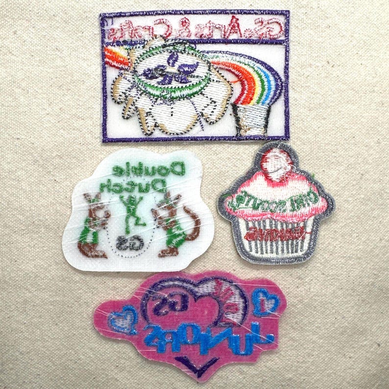 Puede incluir: Cuatro parches bordados sobre un fondo beige. Los parches incluyen un arco&iacute;ris, una magdalena, un coraz&oacute;n y dos personajes de dibujos animados. Los parches tienen varios colores, incluyendo rosa, azul, verde y blanco. Los parches tienen texto.