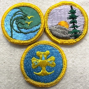 May include: Three embroidered patches with gold trim. One patch depicts a wave with birds, another a mountain scene with a sunset, and the third a gold emblem on a blue background. These patches are likely for a youth organization.