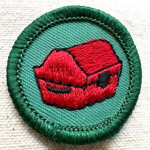 May include: Embroidered patch featuring a red object, possibly a container or box, on a teal background. The object is outlined in black and surrounded by a green border. The patch is circular.