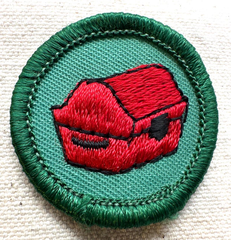 May include: Embroidered patch featuring a red object, possibly a container or box, on a teal background. The object is outlined in black and surrounded by a green border. The patch is circular.