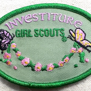 May include: Embroidered oval patch with the words "INVESTITURE GIRL SCOUTS" in green and purple. Features two butterflies and a floral garland in pink and yellow, all outlined in green.