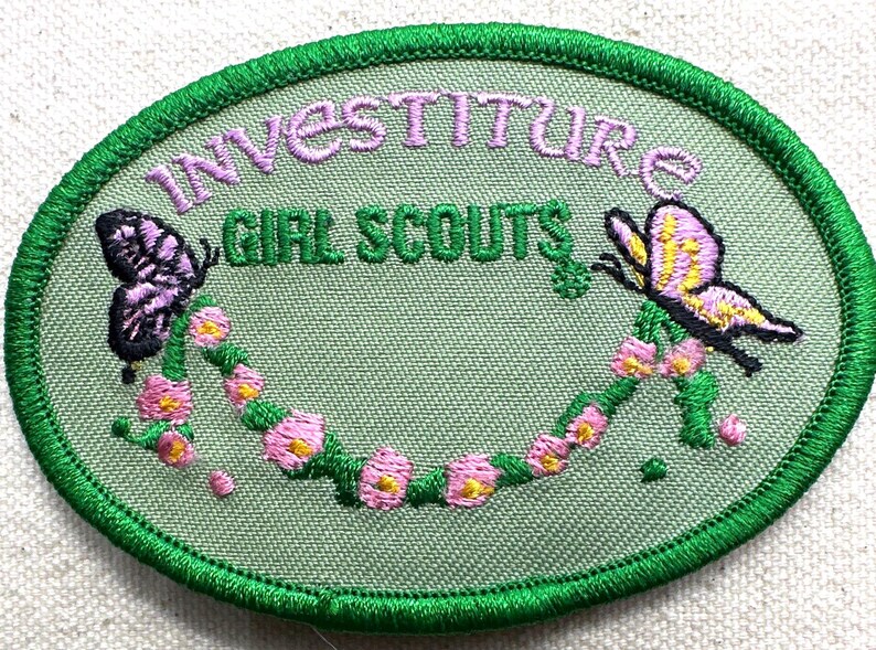 May include: Embroidered oval patch with the words "INVESTITURE GIRL SCOUTS" in green and purple. Features two butterflies and a floral garland in pink and yellow, all outlined in green.