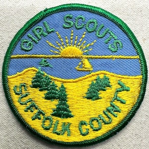 May include: A circular Girl Scouts Suffolk County patch. The patch features a yellow and blue landscape with green trees and a yellow sun. The words "GIRL SCOUTS" and "SUFFOLK COUNTY" are embroidered in green around the edge of the patch.