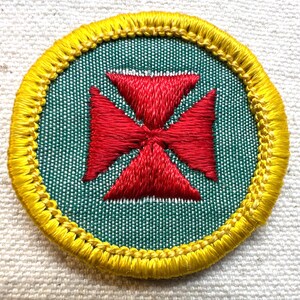 May include: Embroidered patch with a red cross design on a green background, encircled by a yellow border. The patch is round and has a textured appearance, likely made of fabric.