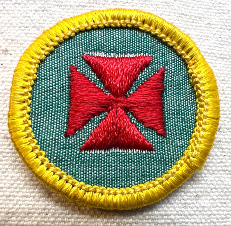 May include: Embroidered patch with a red cross design on a green background, encircled by a yellow border. The patch is round and has a textured appearance, likely made of fabric.