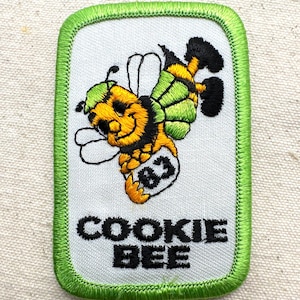 May include: Embroidered patch featuring a cartoon bee with a green hat and wings, and black and yellow stripes. The patch has a white background with the words "COOKIE BEE" in black block letters, and a green border.