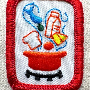 Puede incluir: Parche bordado con borde rojo y fondo blanco. El parche presenta una olla roja con detalles negros y varios alimentos en azul, verde, naranja, blanco y amarillo. El parche es de forma rectangular.