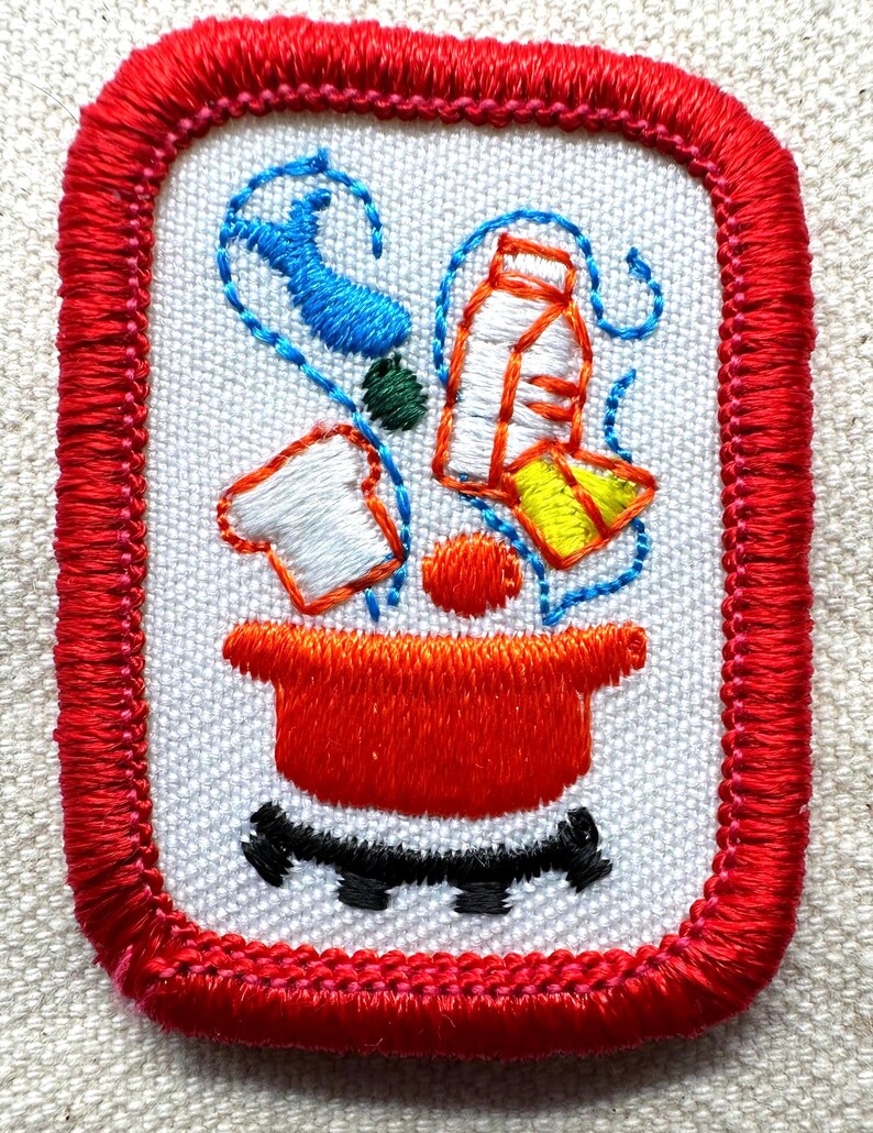 Puede incluir: Parche bordado con borde rojo y fondo blanco. El parche presenta una olla roja con detalles negros y varios alimentos en azul, verde, naranja, blanco y amarillo. El parche es de forma rectangular.