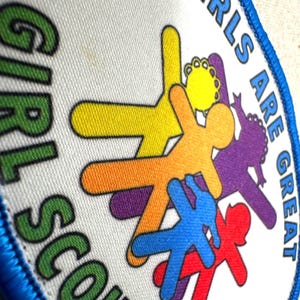 May include: A circular Girl Scouts badge with a blue border. The badge features the words "GIRLS ARE GREAT" and "GIRL SCOUTS" in green and blue. Colourful stick figures in yellow, orange, purple, blue, and red are in the centre.