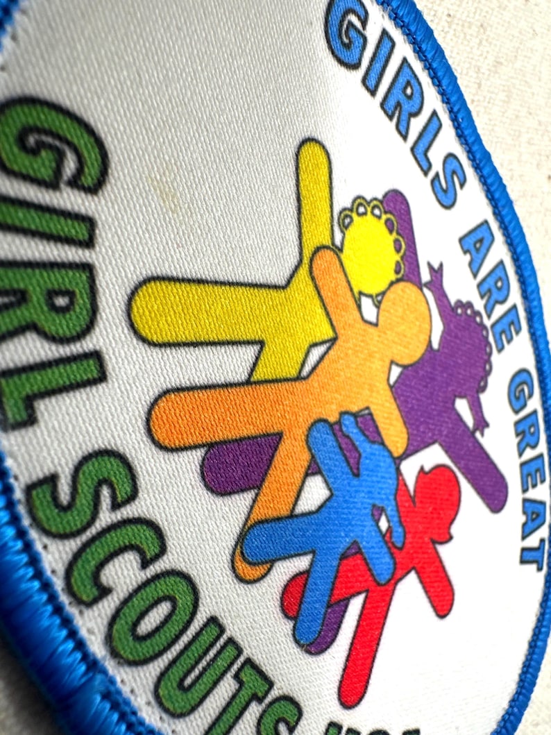 May include: A circular Girl Scouts badge with a blue border. The badge features the words "GIRLS ARE GREAT" and "GIRL SCOUTS" in green and blue. Colourful stick figures in yellow, orange, purple, blue, and red are in the centre.