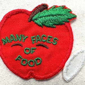 May include: A red apple-shaped patch with green embroidered text that reads "MANY FACES OF FOOD." The apple has a green leaf and a brown stem. A white, oval-shaped patch is next to the apple.