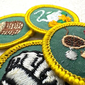 May include: Close-up of several embroidered patches with green backgrounds and yellow borders. The patches feature various designs, including a log, a swan, and a tiger stripe pattern. The patches are likely for a youth organization.