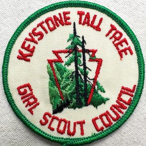 May include: A circular embroidered patch with a cream-colored background and a green border. It features a red arrowhead outline surrounding two black pine trees over green foliage. The text reads KEYSTONE TALL TREE GIRL SCOUT COUNCIL in red.