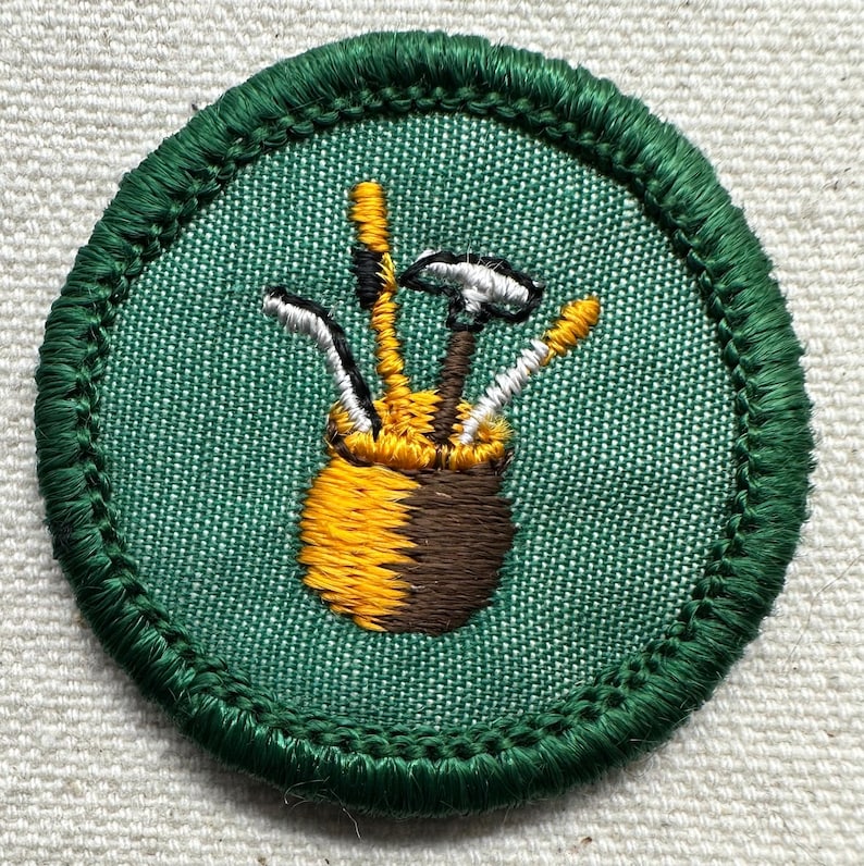 May include: A circular patch with a green border and a green fabric background. The patch features a brown and yellow golf bag with golf clubs. The golf clubs are white, black, and yellow. The patch is likely for a golf-related activity.
