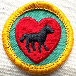 May include: Embroidered patch featuring a black horse silhouette inside a red heart. The heart is set against a green background, framed by a yellow border with gold stitching. A unique, handcrafted design.
