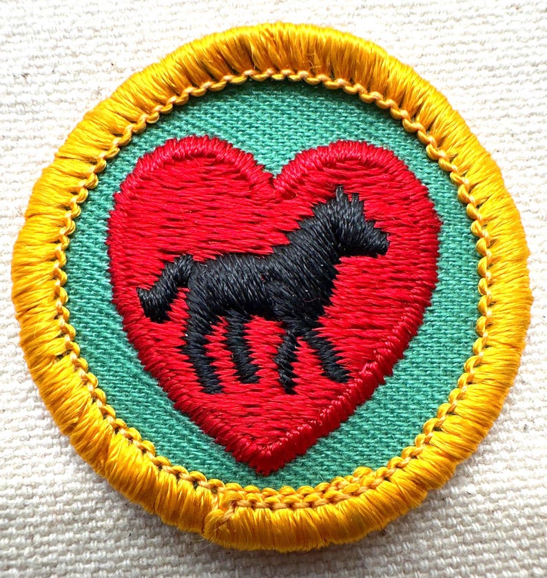 May include: Embroidered patch featuring a black horse silhouette inside a red heart. The heart is set against a green background, framed by a yellow border with gold stitching. A unique, handcrafted design.