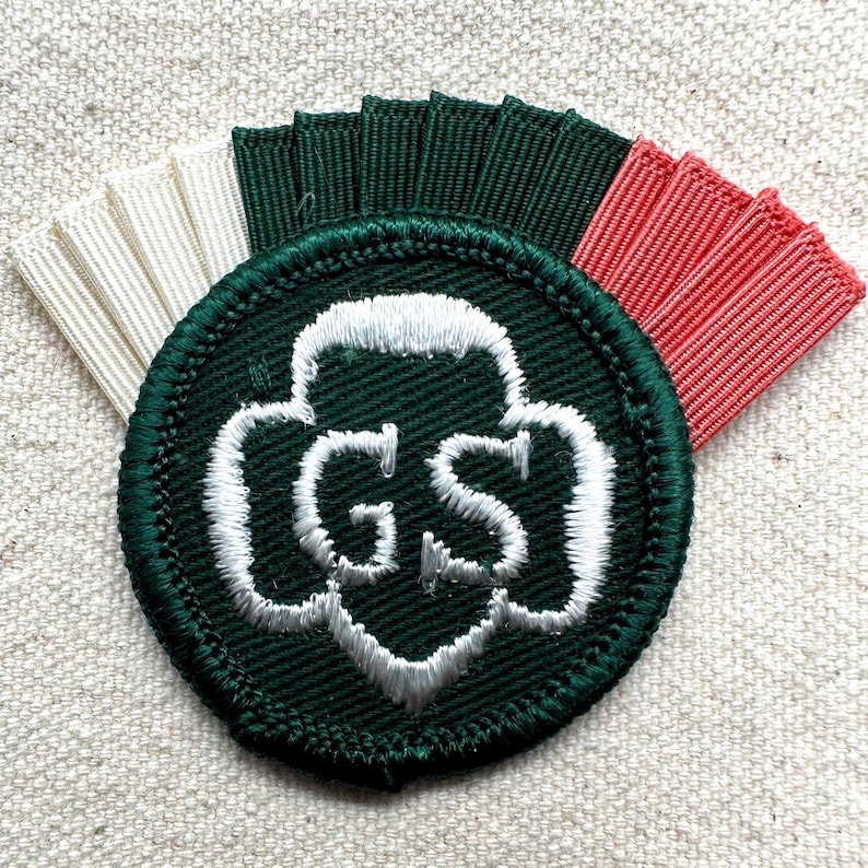 May include: A circular green embroidered patch with the letters "GS" in white, surrounded by a white outline. Ribbons in white, green, and red fan out from the patch. The patch is likely a Girl Scout emblem.