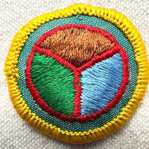 May include: Embroidered patch with a circular design. The patch features a segmented interior with sections of brown, green, and blue, outlined in red. The outer edge is green, and the border is yellow. The patch is likely for a uniform or craft project.