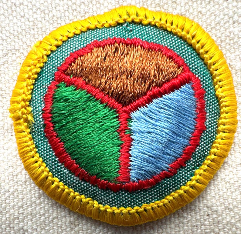 May include: Embroidered patch with a circular design. The patch features a segmented interior with sections of brown, green, and blue, outlined in red. The outer edge is green, and the border is yellow. The patch is likely for a uniform or craft project.