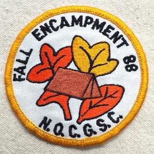 May include: A circular patch with a white background, featuring a brown tent, orange and yellow leaves, and the text "FALL ENCAMPMENT 88 N.Q.C.G.S.C." in black. The patch has a yellow border.