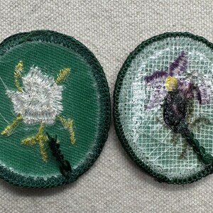 Pode incluir: Dois patches bordados com desenhos florais. Um apresenta uma flor branca com detalhes dourados sobre um fundo verde. O outro mostra uma flor roxa e amarela sobre um fundo de malha branca, ambos com bordas verdes.