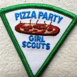 Puede incluir: Un parche triangular con fondo blanco y borde cosido verde. El parche presenta las palabras "PIZZA PARTY" y "GIRL SCOUTS" en azul, con una pizza bordada en el centro.