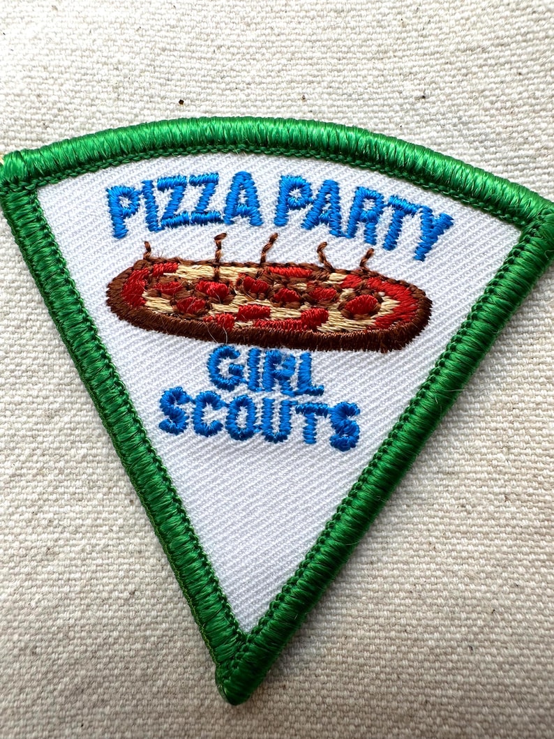Puede incluir: Un parche triangular con fondo blanco y borde cosido verde. El parche presenta las palabras "PIZZA PARTY" y "GIRL SCOUTS" en azul, con una pizza bordada en el centro.