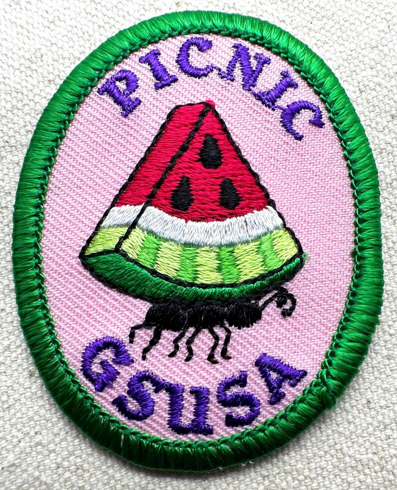 May include: Embroidered patch with a pink oval background and a green border. The patch features a watermelon slice with black seeds, a black ant, and the words "PICNIC" and "GSUSA" in purple.