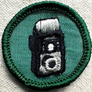 May include: Embroidered patch featuring a black and white camera design on a teal background. The patch has a green border and is likely for a scout or hobbyist. The camera design is detailed with white and black thread.